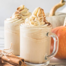 Two glasses of iced pumpkin spice latte with whipped cream, sprinkled with pumpkin spice. You can see cinnamon and pumpkins in the background.